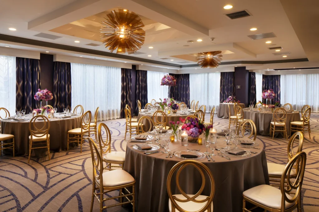 Elegant banquet hall among top event venues in Costa Mesa, featuring round tables with gray tablecloths, gold chairs, floral centerpieces, and place settings. Large windows with sheer curtains and gold chandeliers enhance the sophisticated décor.