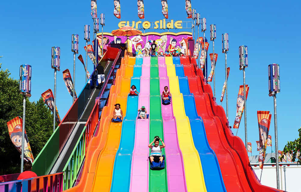 Children and adults slide down colorful lanes of a giant Euro Slide amusement park ride under a clear blue sky—a highlight among Costa Mesa events—flanked by decorative flags and twinkling lights on either side.