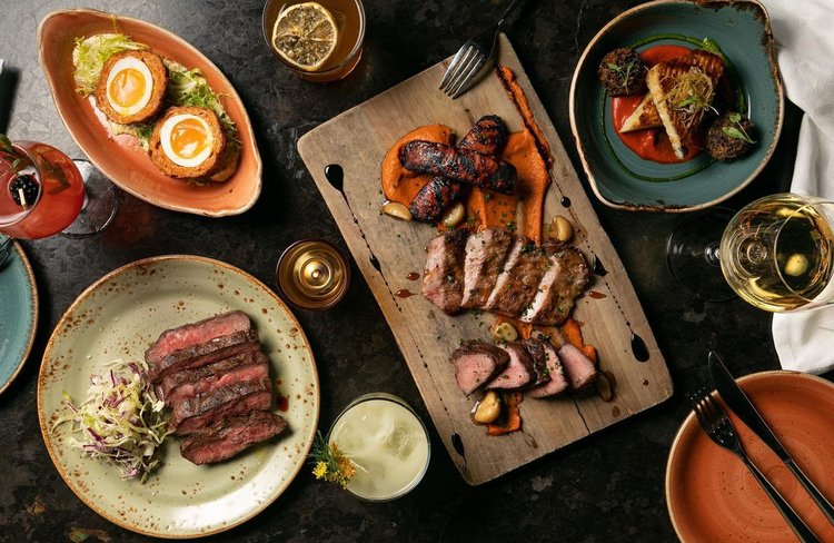 A table set with various gourmet dishes, including sliced steak, grilled sausages, vegetables, salad, and cocktails, arranged artfully on rustic plates and a wooden board—reflecting the creative spirit of Costa Mesa City of the Arts.