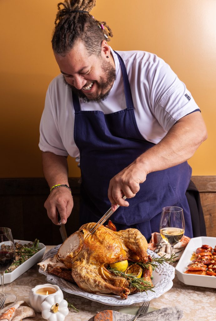 A smiling man in a blue apron carves a roasted turkey at a table set with side dishes and wine glasses, against a warm orange background.