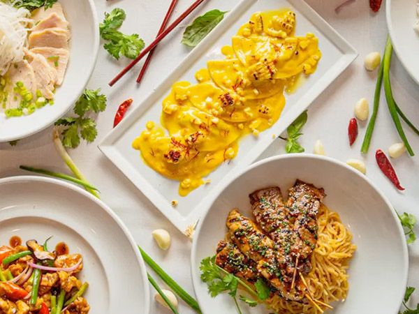 A table set with various Asian dishes, including yellow curry on a rectangular plate, grilled meat on noodles, sliced chicken, and stir-fried vegetables, surrounded by fresh herbs, chilies, and garlic.