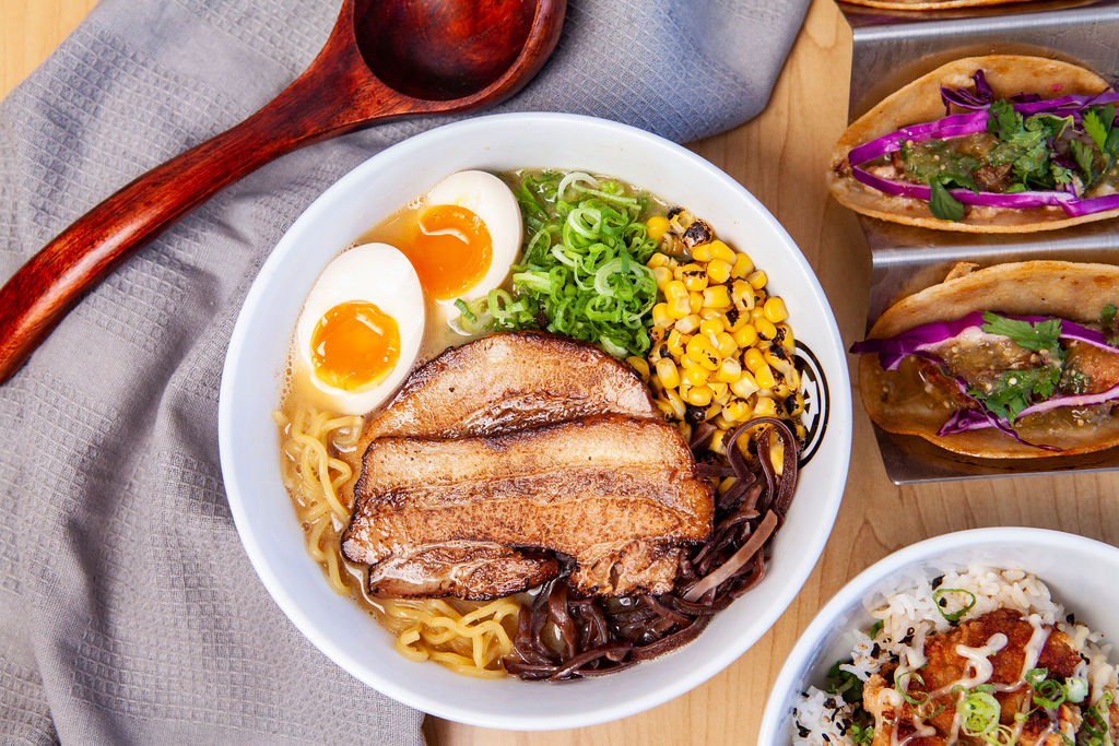 A bowl of ramen topped with two soft-boiled egg halves, sliced pork belly, corn, green onions, and mushrooms sits on a table beside tacos and a rice dish—a delicious highlight among Costa Mesa attractions.