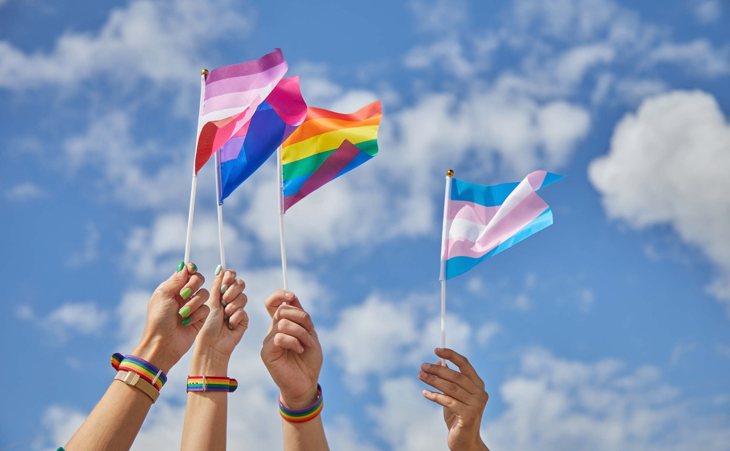Four hands hold up different LGBTQ+ pride flags—lesbian, bisexual, rainbow, and transgender—against a blue sky with clouds. Rainbow bracelets on each wrist show pride and support, perfect for a Costa Mesa travel blog celebrating diversity.