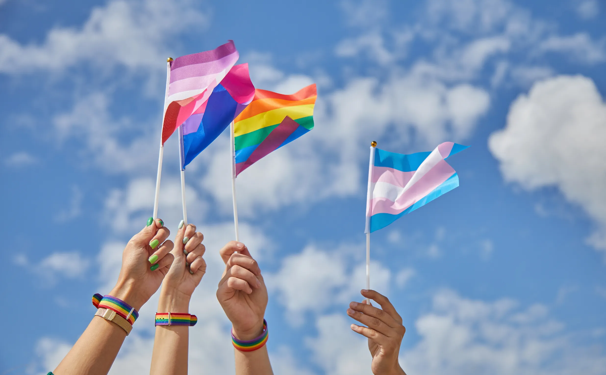Four hands hold up different LGBTQ+ pride flags—lesbian, bisexual, rainbow, and transgender—against a blue sky with clouds. Rainbow bracelets on each wrist show pride and support, perfect for a Costa Mesa travel blog celebrating diversity.