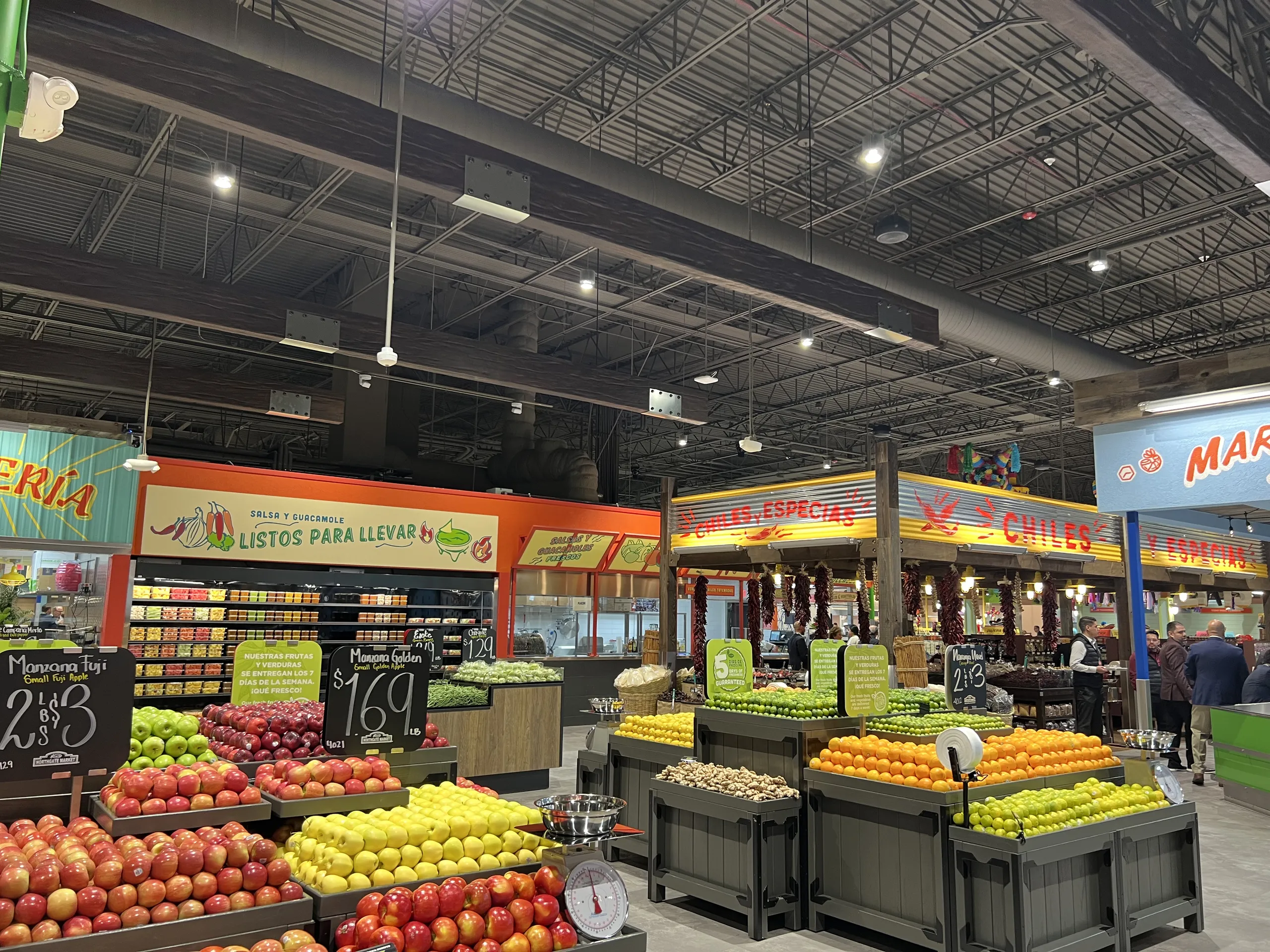 A brightly lit grocery store produce section displays apples, oranges, and lemons. Colorful signs in Spanish hang above, and sections for meats and other goods are visible in the background—an inviting scene you might find highlighted on a Costa Mesa travel blog.