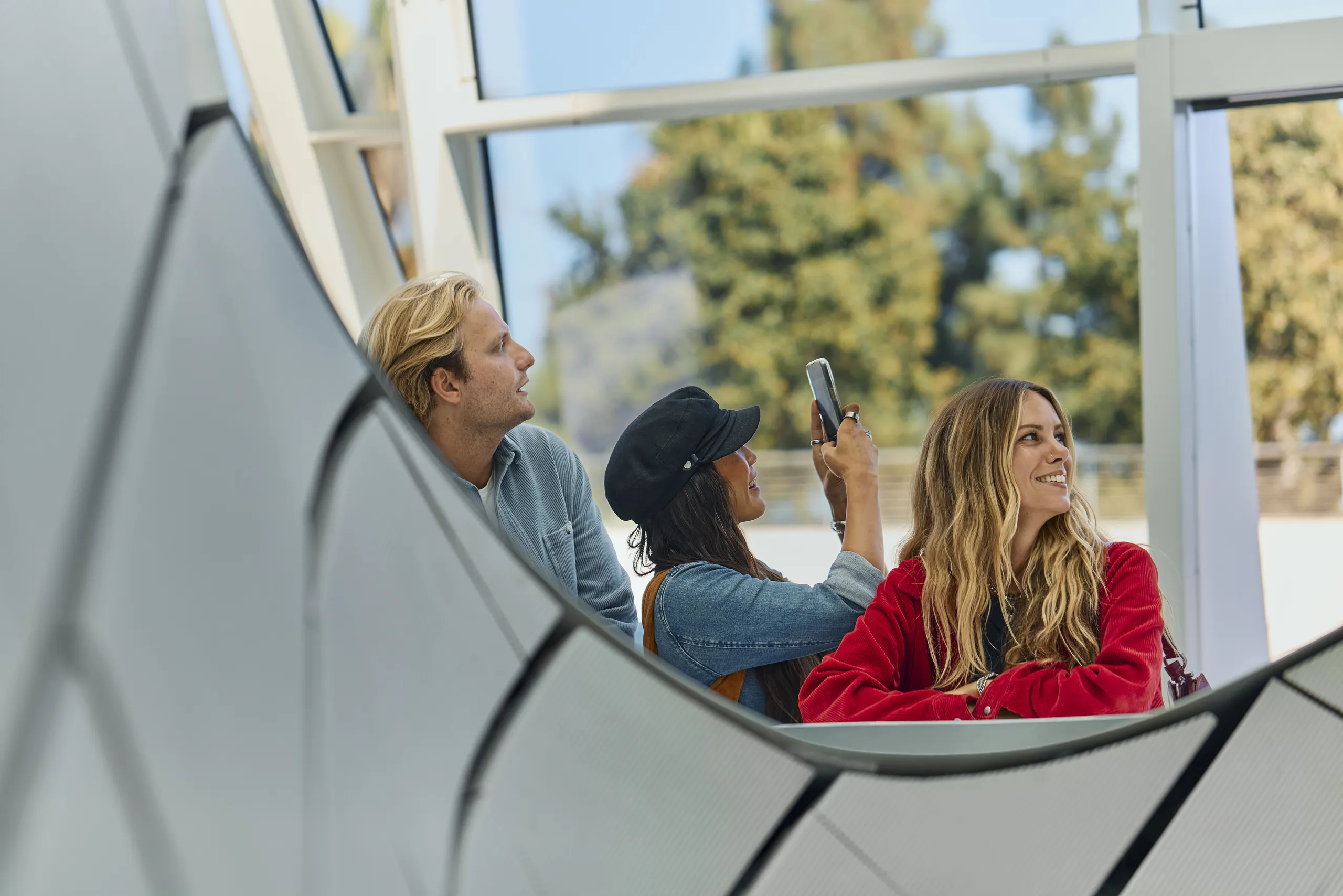 Three people stand indoors near large windows, perhaps capturing memories for a Costa Mesa travel blog. One person in a black hat takes a photo with a phone, while the other two, a man and a woman, smile and look off in different directions. Lush trees are visible outside.