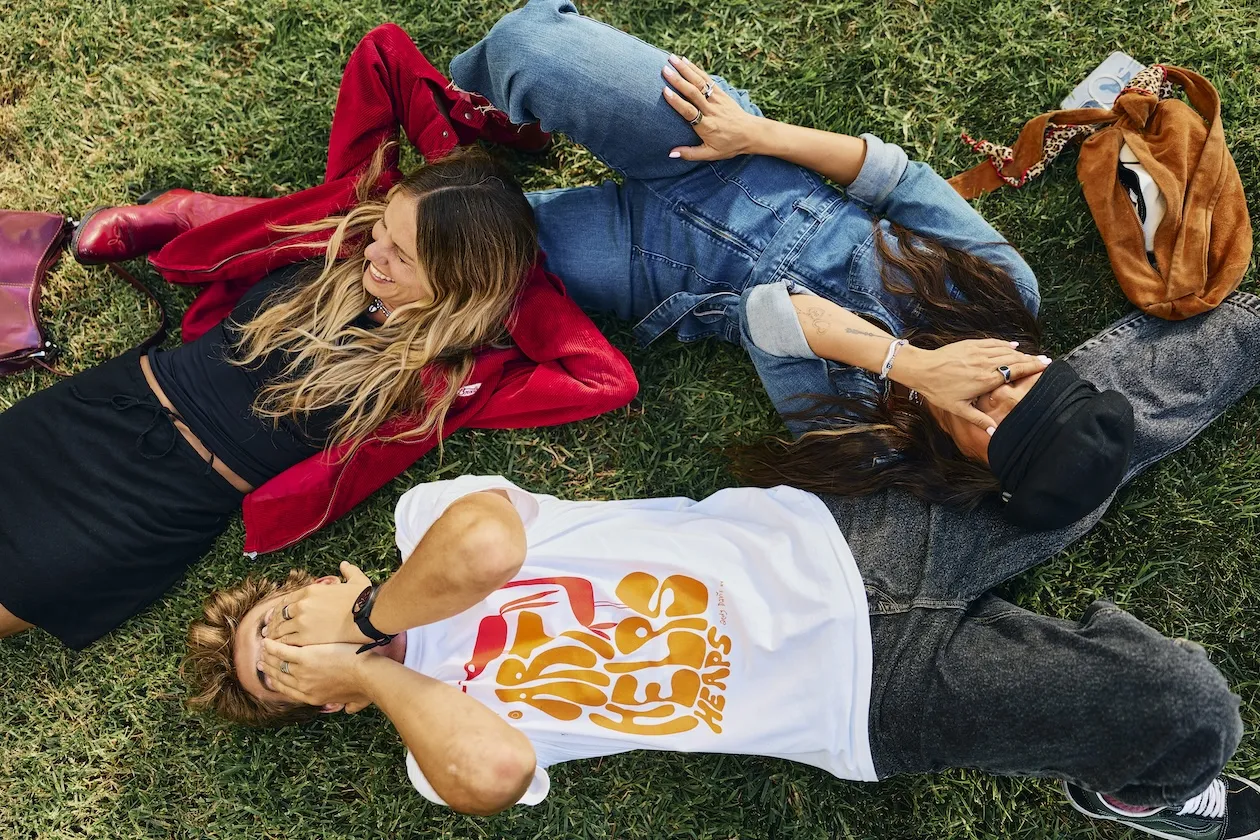 Three friends lying on grass outdoors, laughing and covering their eyes playfully. With backpacks and a purse nearby, it’s a carefree moment perfect for any Costa Mesa travel blog.