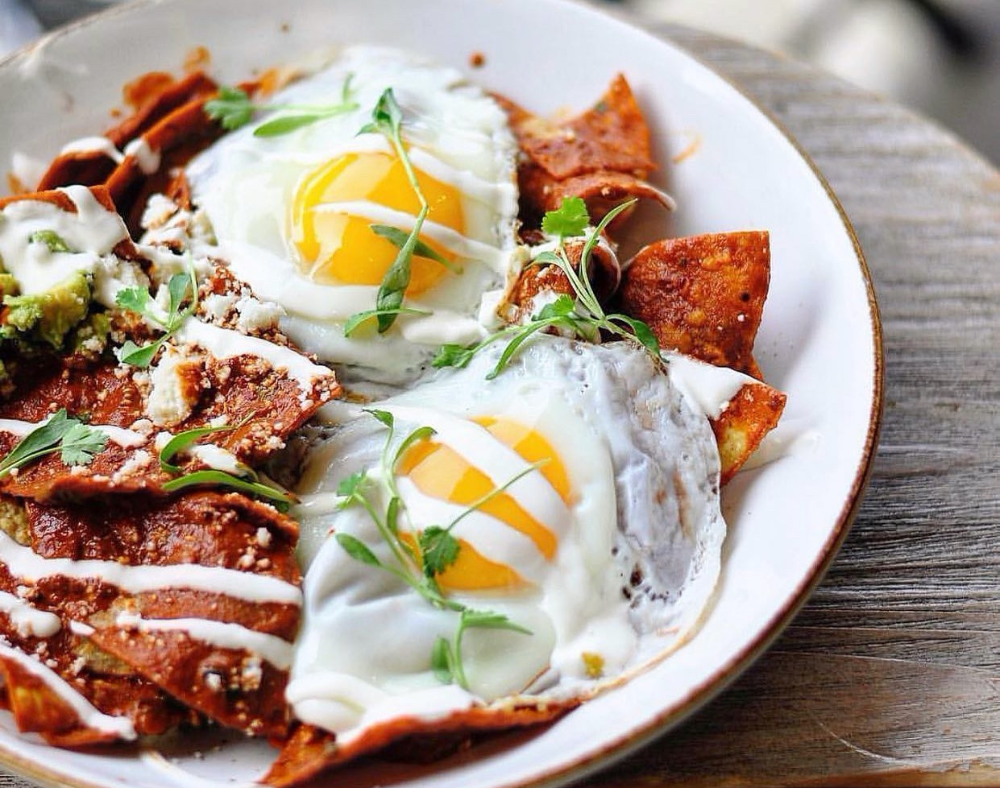 A plate of chilaquiles topped with two sunny-side-up eggs, fresh herbs, crema, and crumbled cheese, served on a rustic wooden table—perfect for featuring on any Costa Mesa travel blog.