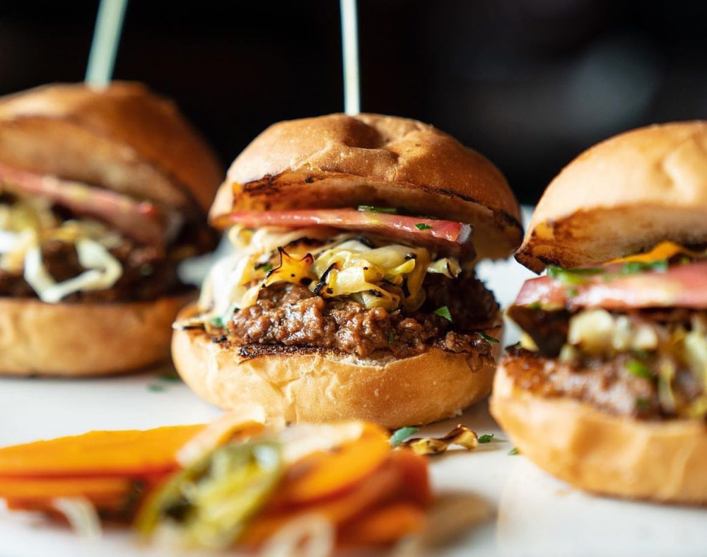 Three juicy burgers topped with grilled onions, melted cheese, and tomato slices sit on a white surface, with a colorful side of sliced pickles and vegetables—a perfect feature for any Costa Mesa travel blog craving local flavors.