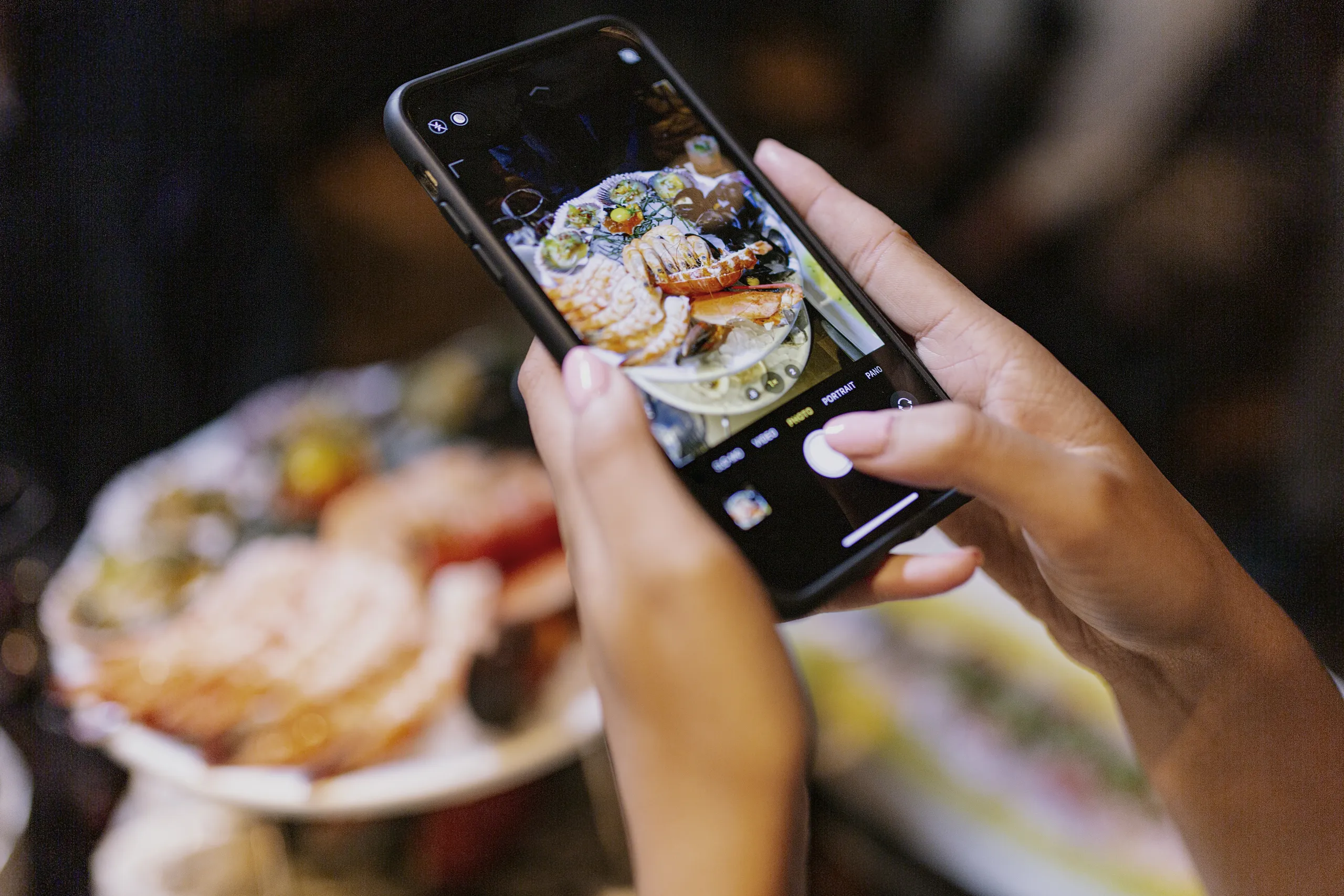 A close-up shot shows a person's hands holding a smartphone, capturing a photo of a seafood platter with lobster and shrimp, which is also visible in the slightly blurred background. The phone's camera interface is visible on the screen, perfect for featuring in a Costa Mesa travel blog.