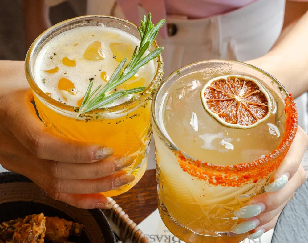 Two hands holding cocktails; one adorned with rosemary and ice, the other with a chili rim and dried citrus. Both drinks, in textured glasses, are viewed from above—a perfect shot for any Costa Mesa travel blog.