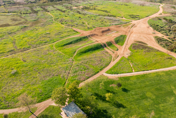 Aerial view of a park with winding dirt paths crossing green fields and patches of grass, trees scattered throughout, and a small structure in the bottom left corner—perfect for a Costa Mesa travel blog feature.