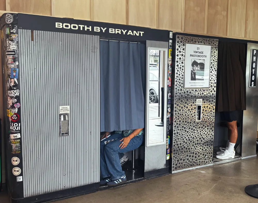 Two vintage photo booths stand side by side. One has people partially visible behind a blue curtain; the other shows a person’s legs below a dark curtain. Both booths have stickers and signage on their exterior.