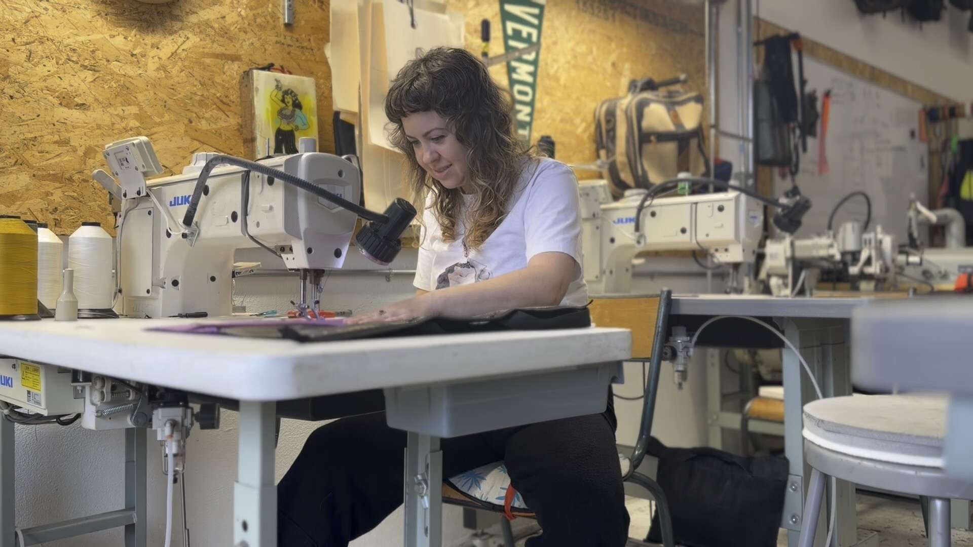 A woman sits at a sewing machine in a workshop, focused on working with fabric. Various sewing tools and equipment are visible around her on tables and the walls.