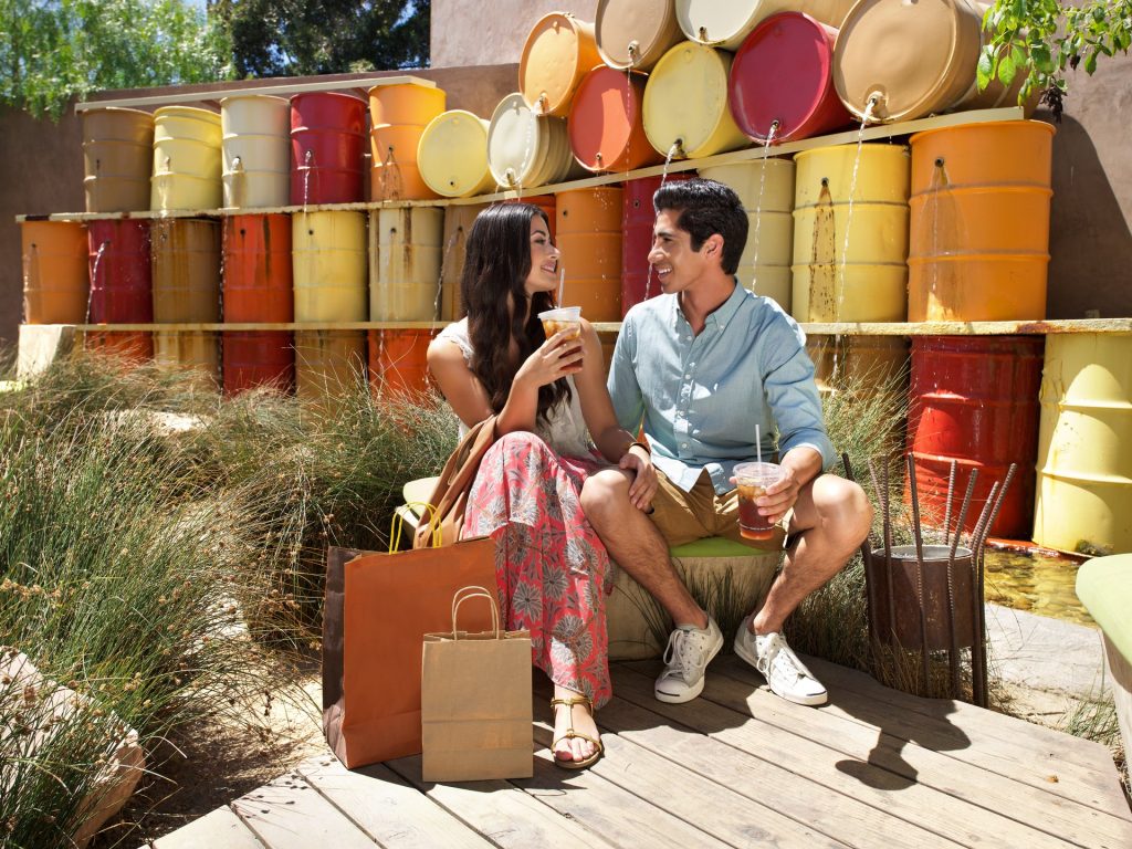 A young couple sits on a bench outdoors, smiling at each other and holding drinks. Shopping bags are by their feet, and colorful stacked barrels form a vibrant backdrop. Grasses grow around the wooden deck where they sit.
