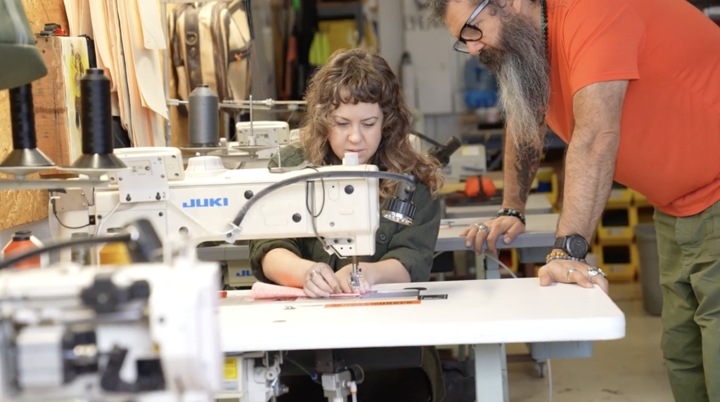 A woman uses a sewing machine at a worktable while a man with a beard and glasses stands beside her, observing. They are in a workshop with sewing equipment and supplies visible in the background.