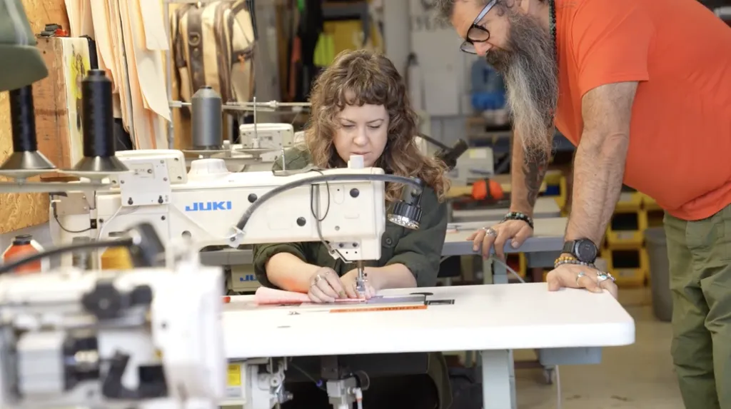 A woman uses a sewing machine at a worktable while a man with a beard and glasses stands beside her, observing. They are in a workshop with sewing equipment and supplies visible in the background.