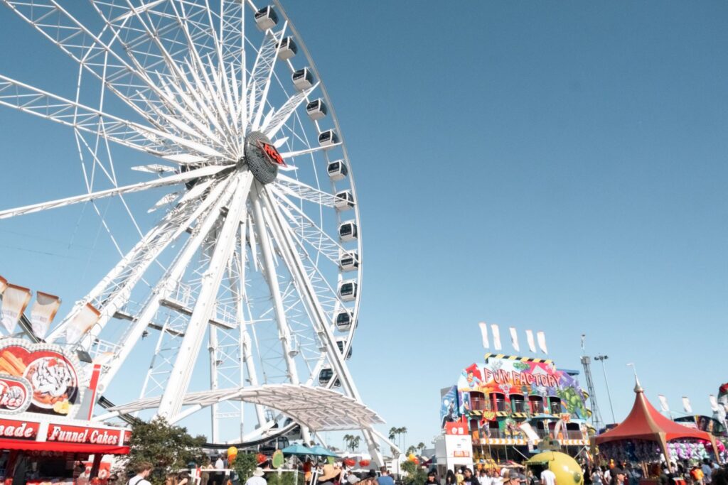 A large Ferris wheel towers over the busy OC Fair Costa Mesa fairground, filled with colorful booths, food stands, and crowds of people under a clear blue sky.
