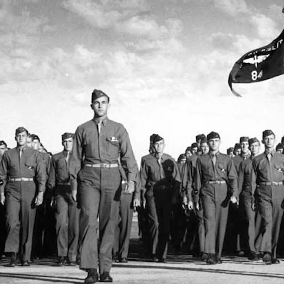 A group of uniformed soldiers marches in formation outdoors under a cloudy sky, with a flag waving in the upper right corner—a striking scene reflecting moments found in the history of Costa Mesa. One soldier leads the group front and center.