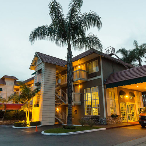 A small hotel building with exterior staircases, large windows, and palm trees in front—near the OC Fair Costa Mesa—glows with warm yellow lights at dusk. A car is parked by the entrance, welcoming visitors.