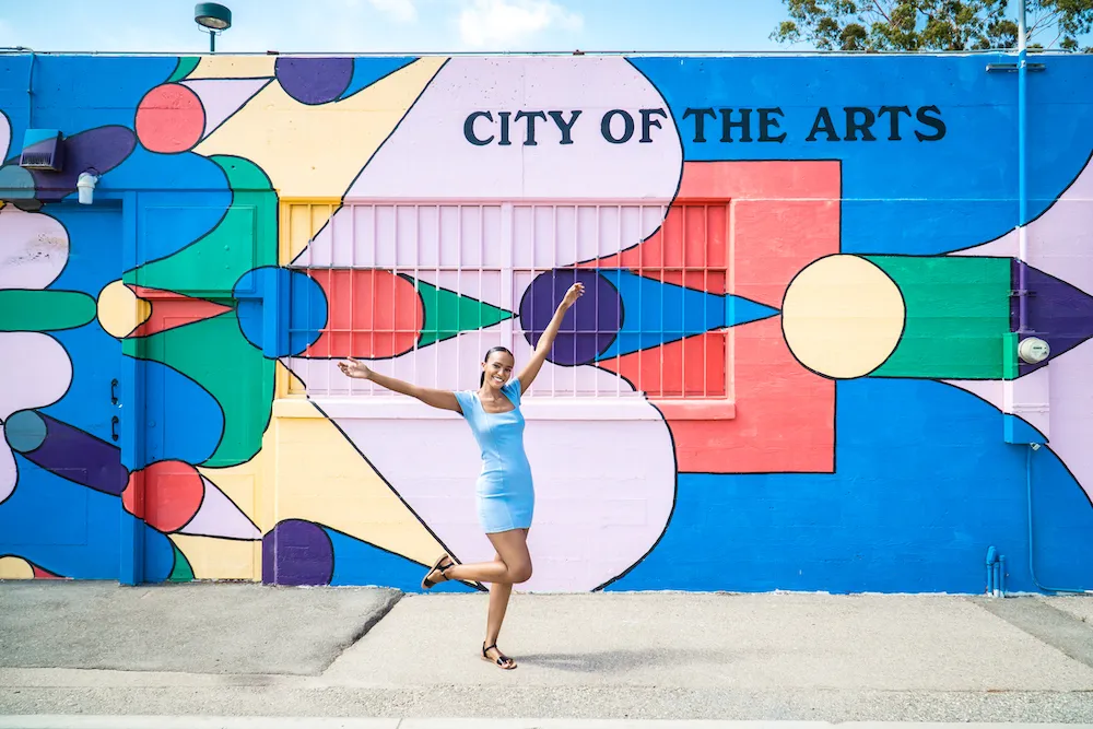 A woman in a light blue dress poses joyfully in front of a colorful geometric mural that reads CITY OF THE ARTS. The mural features bold shapes and vibrant colors, and the woman stands on one leg with her arms raised.