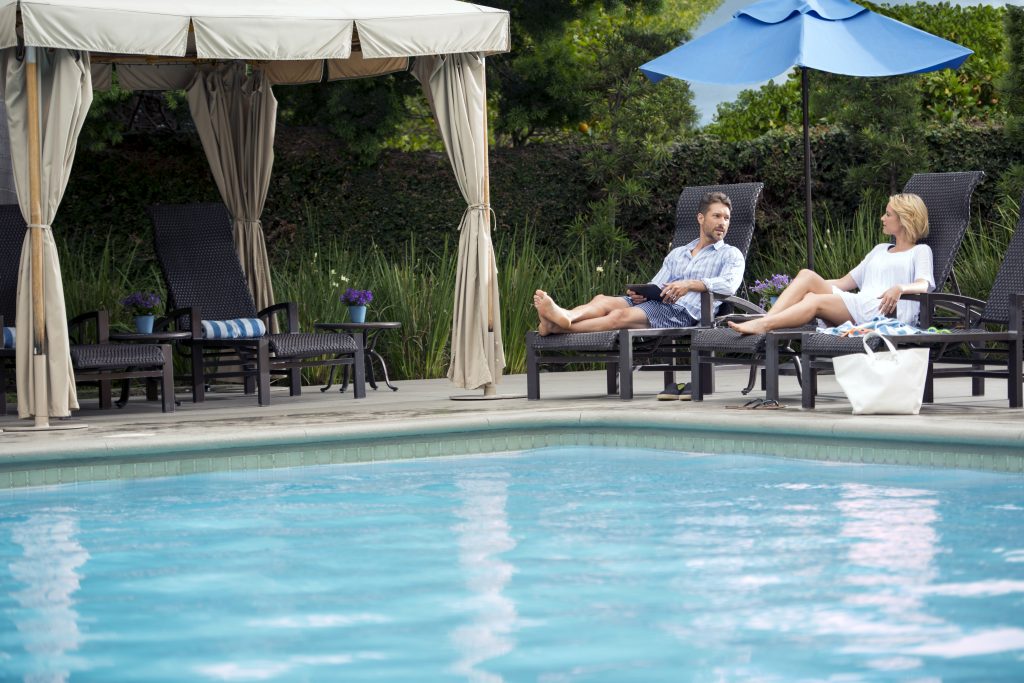 A man and woman relax on lounge chairs by a pool under a canopy and blue umbrella, with greenery in the background and a white tote bag on the ground nearby.