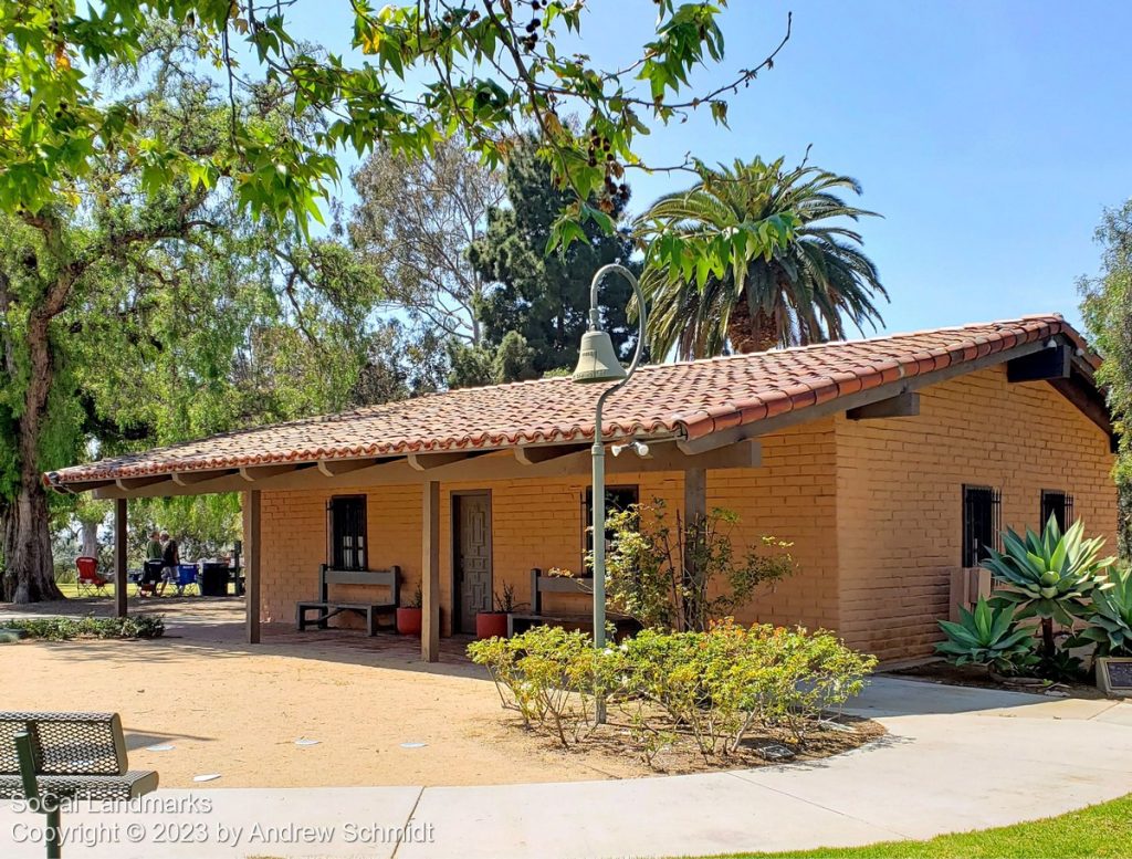 A one-story adobe building with a red tiled roof and wooden beams sits surrounded by trees and plants under a clear sky. A shaded outdoor area with people is visible on the left.
