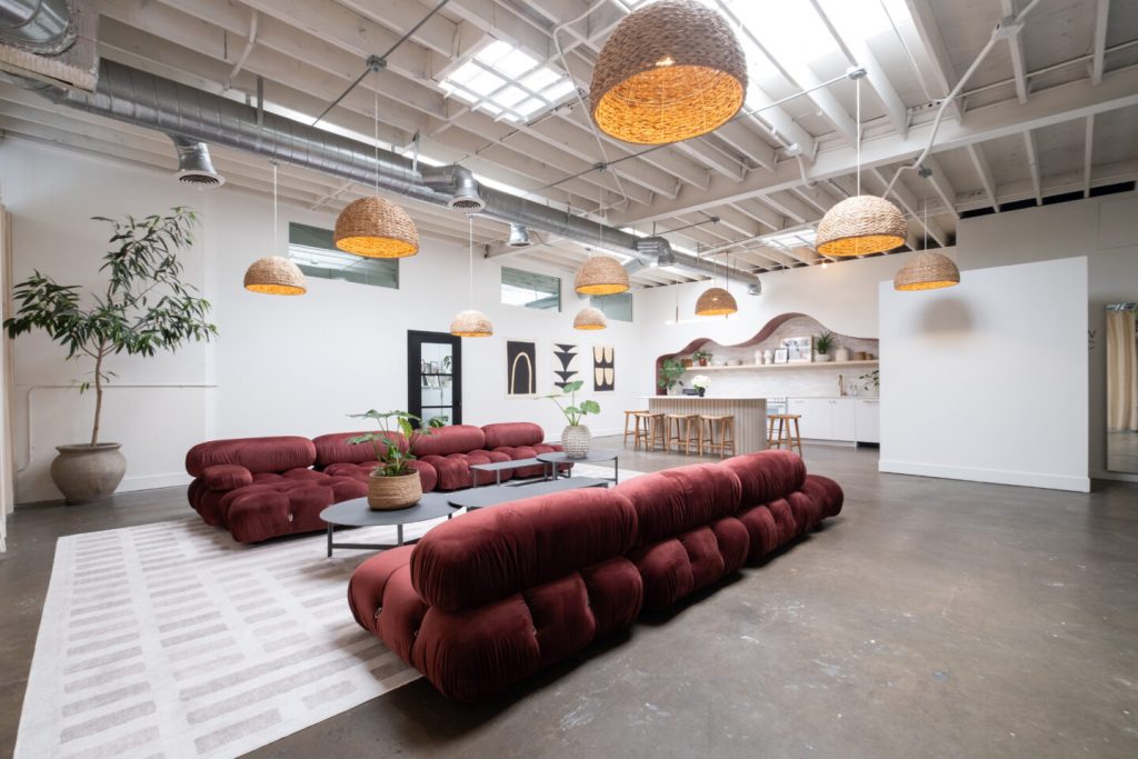 A modern, spacious lounge with maroon tufted sofas, woven pendant lights, potted plants, and a kitchen area with barstools. Exposed ceiling beams and large windows create an airy, inviting atmosphere.