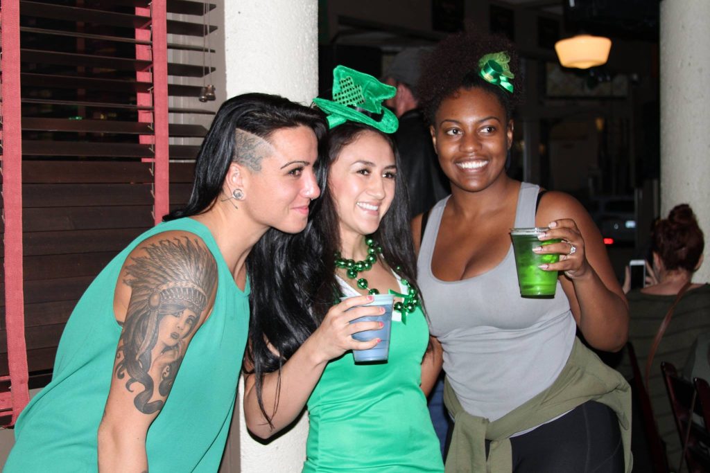 Three women smiling and holding drinks, dressed in green attire and accessories, celebrating indoors. One woman has a visible tattoo on her arm. The scene suggests a festive occasion, likely St. Patricks Day.