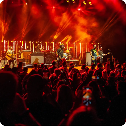 A band performs on a brightly lit stage with orange and yellow lights at the OC Fair Costa Mesa, as a large crowd watches and takes photos with their phones during the lively concert.