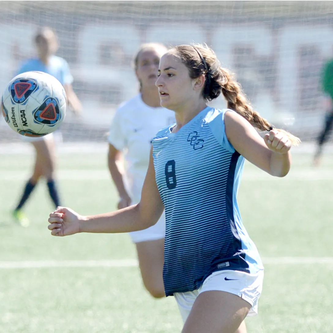 A female soccer player in a blue and white jersey prepares to control a ball mid-air during a sports event in Costa Mesa, with other players and a goal net visible in the background.