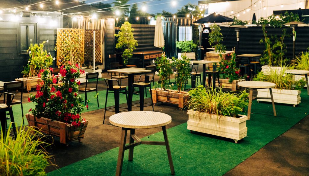 Outdoor patio with green artificial turf, scattered tables and chairs, potted plants, string lights overhead, and a cozy, inviting atmosphere in the evening.