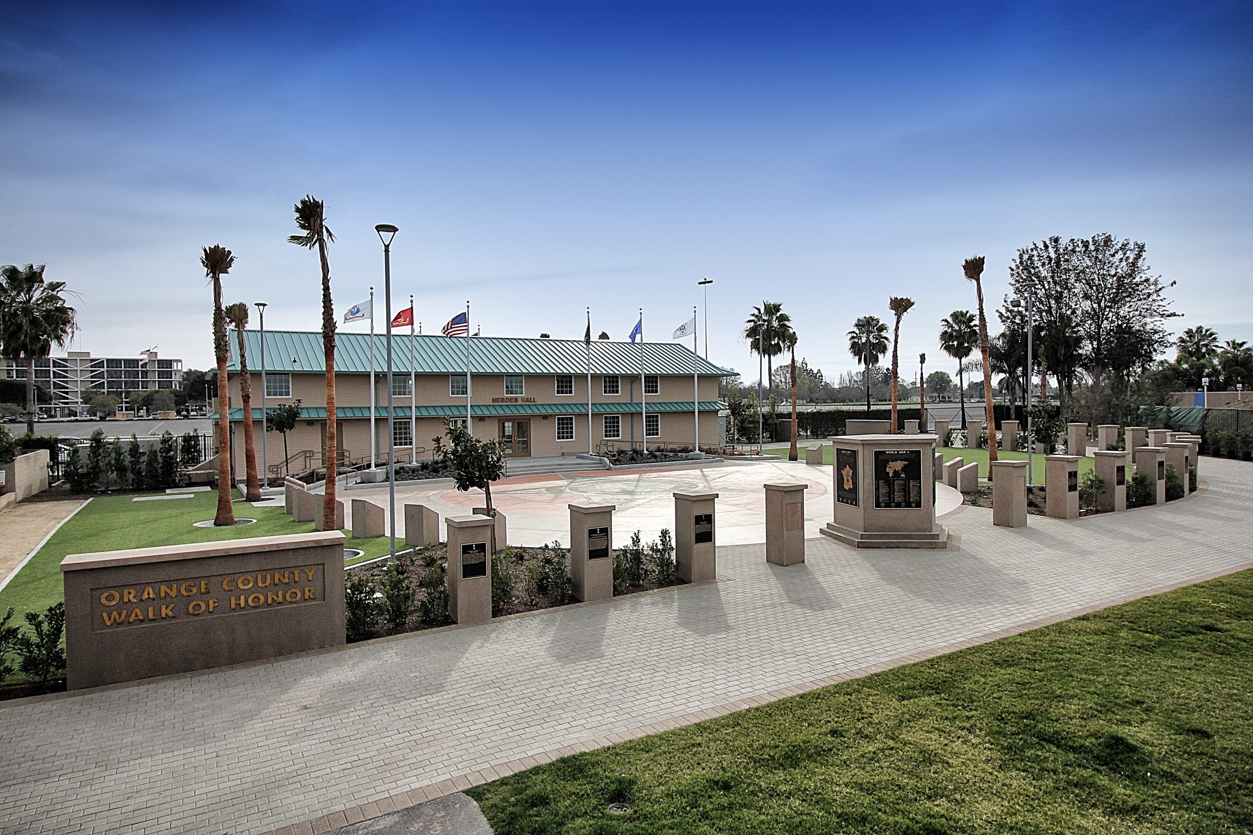 A paved walkway leads to a building with a green roof, surrounded by palm trees and flagpoles with flags—an ideal spot for any Costa Mesa travel blog exploring the Orange County Walk of Honor memorial area lined with stone pillars.