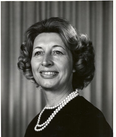 Black and white portrait of a smiling woman with short, curled hair, wearing a dark top and a double-strand pearl necklace. She is posed in front of a plain, softly striped background.