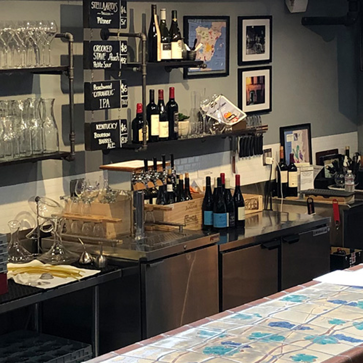A bar counter with wine bottles, glasses, and a chalkboard menu on the wall. Shelves hold glassware and framed pictures hang above the counter. A colorful tiled surface is in the foreground.