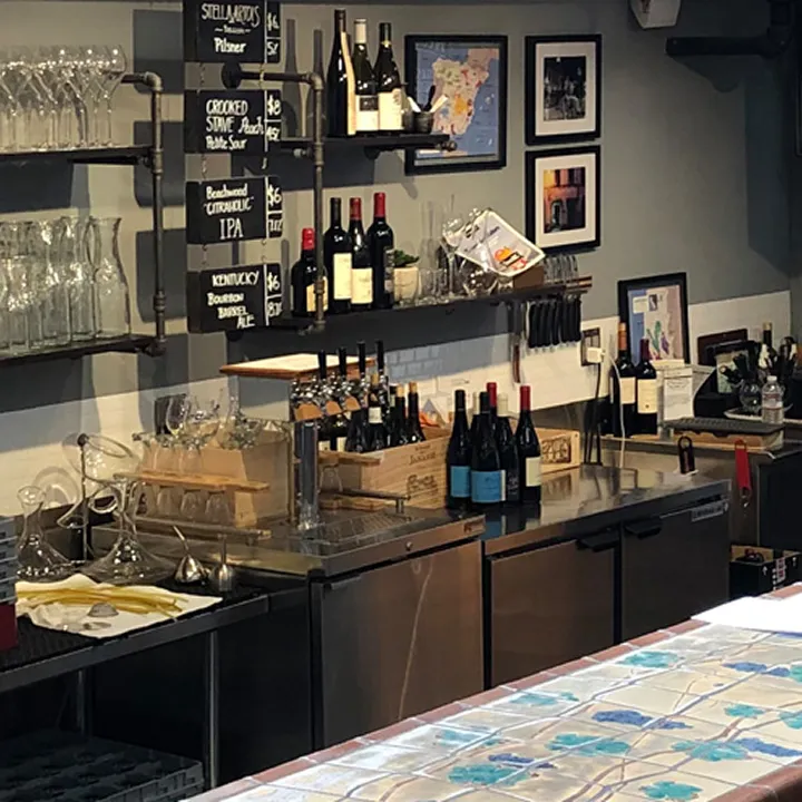 A bar counter with wine bottles, glasses, and a chalkboard menu on the wall. Shelves hold glassware and framed pictures hang above the counter. A colorful tiled surface is in the foreground.