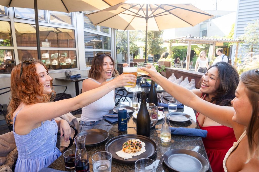 Four women sit at an outdoor restaurant table in Costa Mesa, smiling and raising their drinks in a toast. Plates of food and drinks are on the table, as large umbrellas provide shade—a perfect moment for a Costa Mesa travel blog.