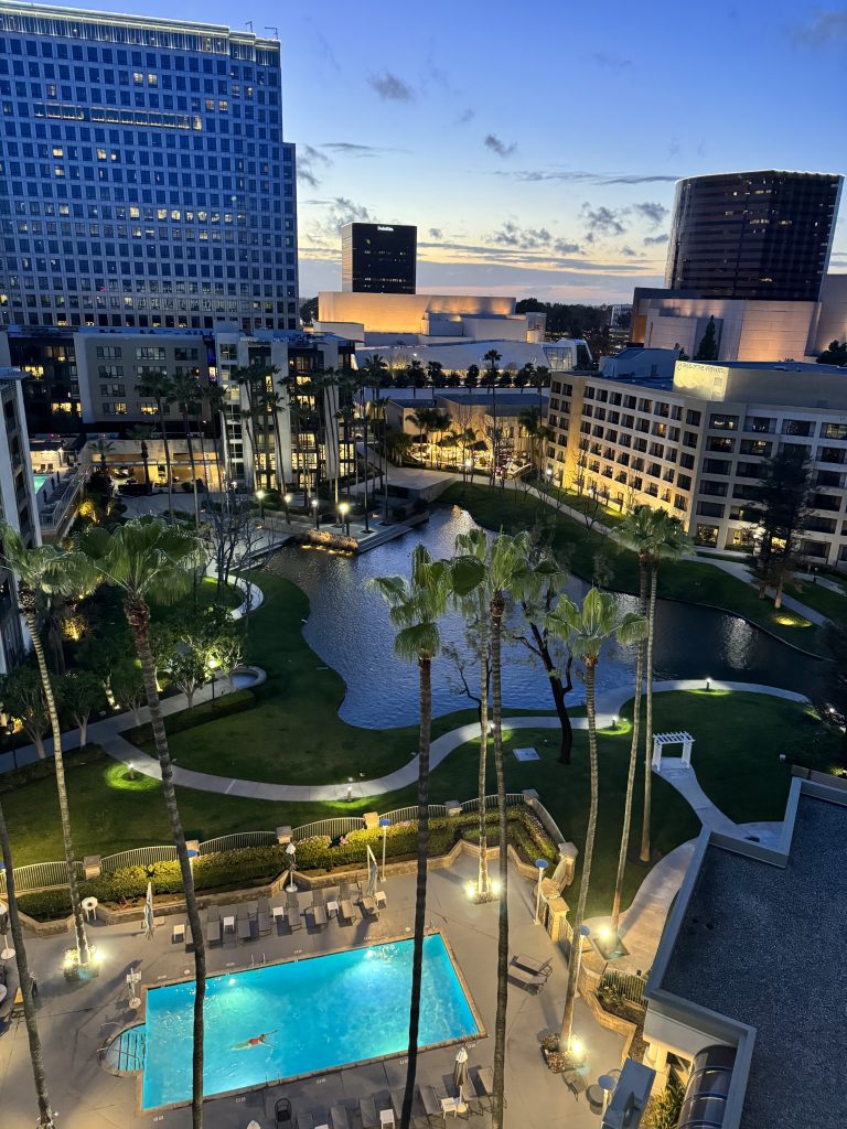 A high-angle view of a cityscape at dusk shows tall buildings, a lit swimming pool, palm trees, and a pond surrounded by greenery, with the sky glowing orange and blue as the sun sets in the distance.