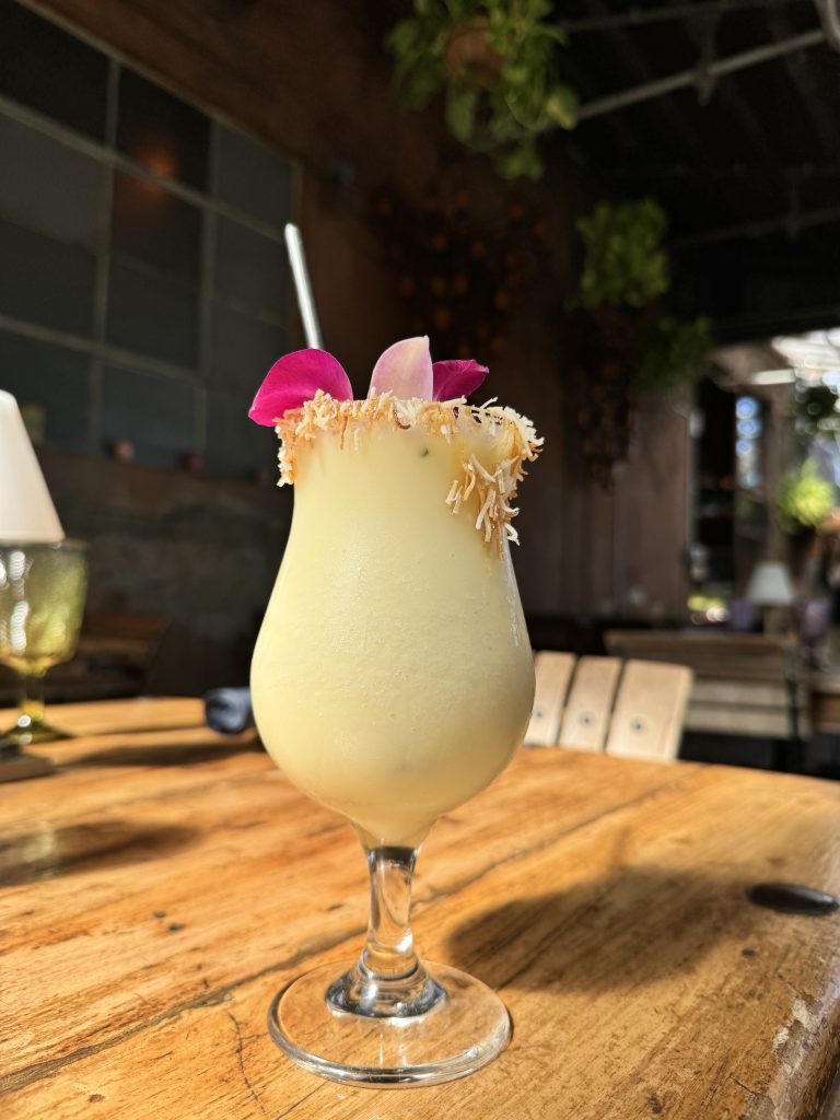 A frosty yellow cocktail in a curved glass, garnished with a pink flower and toasted coconut on the rim, sits on a wooden table in a sunlit, rustic cafe setting with greenery in the background.
