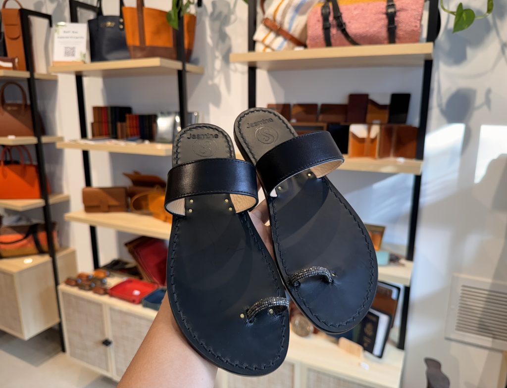 A hand holds up a pair of black leather sandals with toe loops inside a store, with shelves displaying various bags, wallets, and accessories in the background.