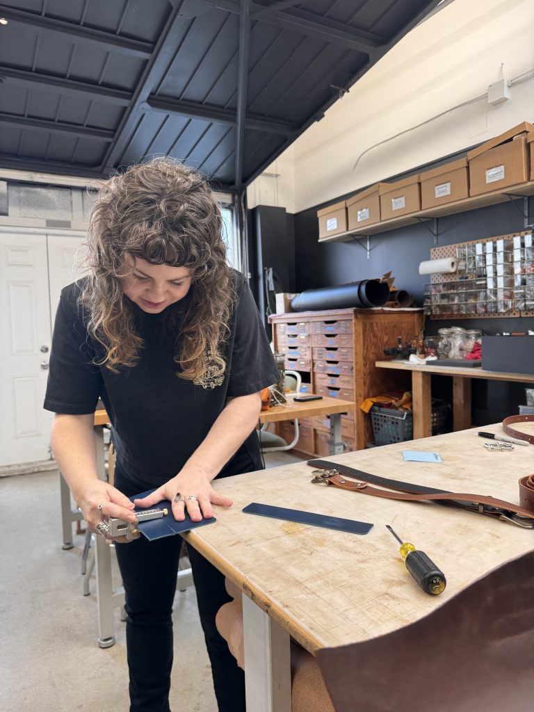 A person with curly hair works on a piece of leather at a wooden table in a workshop, surrounded by tools and materials, including a screwdriver and leather straps.
