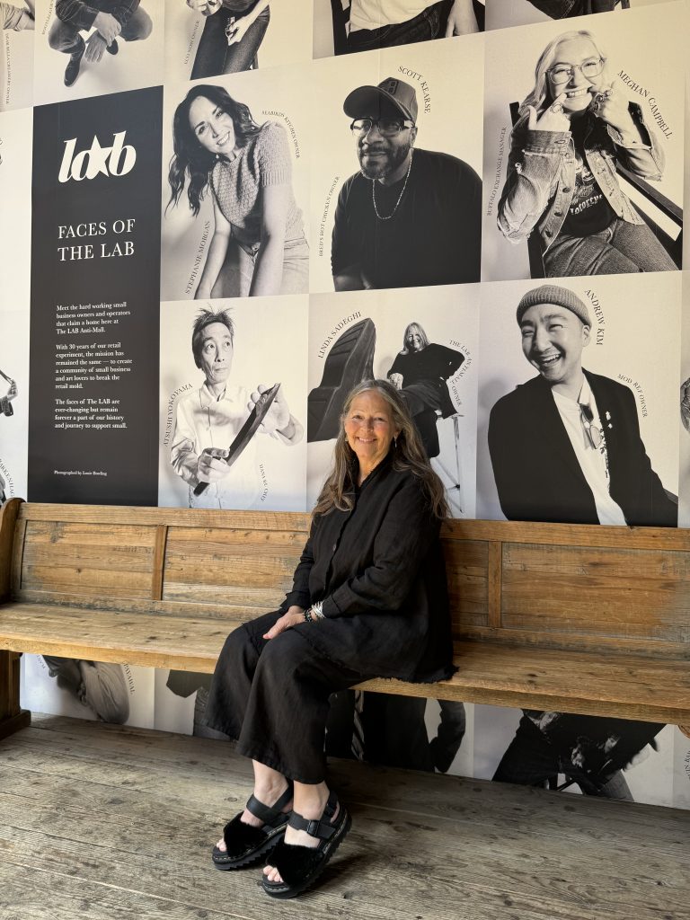 A woman with long gray hair, dressed in black, sits on a wooden bench in front of a wall covered with black-and-white portraits and the text “Faces of the Lab.” Bright natural light fills the space.