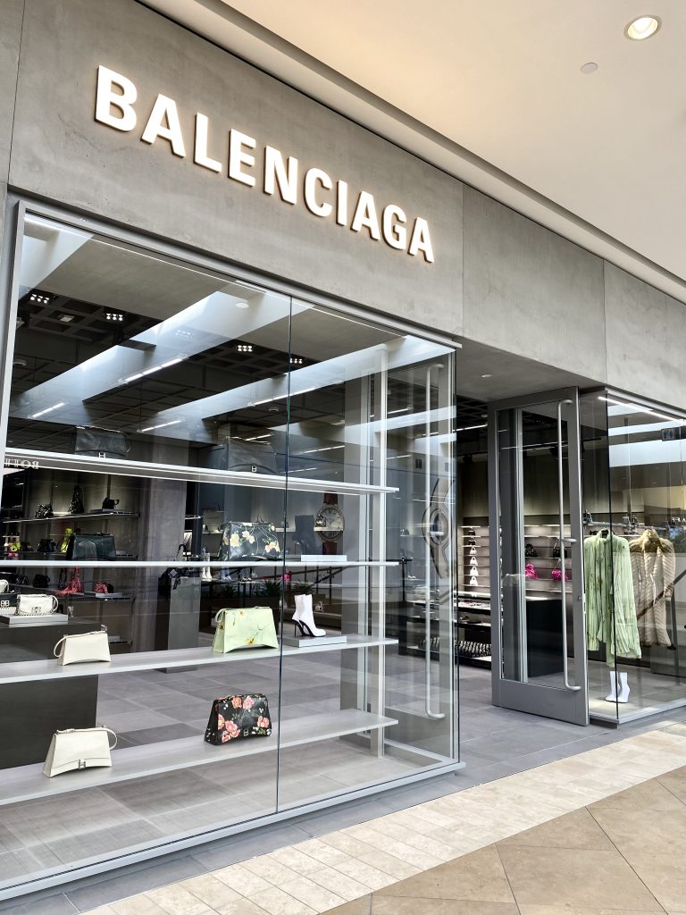 Balenciaga store exterior with large glass windows displaying handbags and accessories; the brand name BALENCIAGA is illuminated above the entrance.