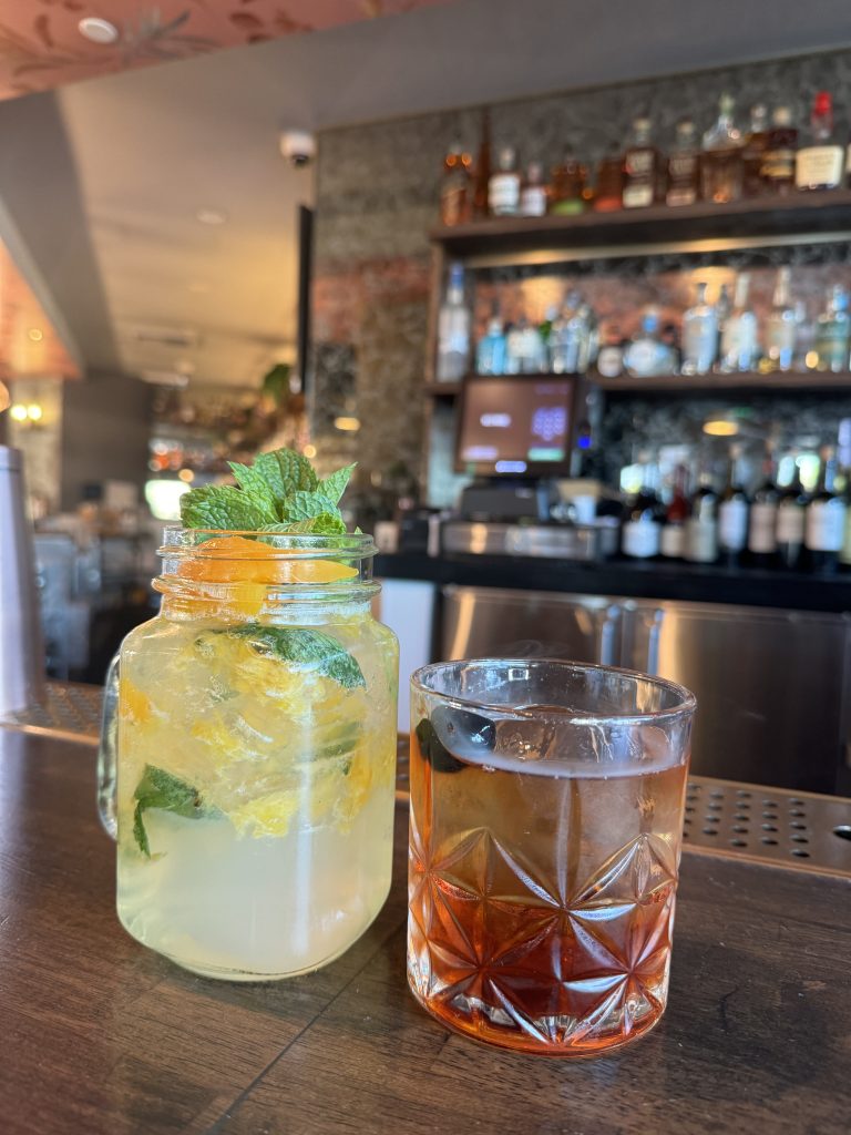 Two cocktails on a bar: one in a mason jar with lemon slices, mint, and ice; the other in a short glass with a dark amber drink and a cherry. Shelves with bottles and a TV are in the background.