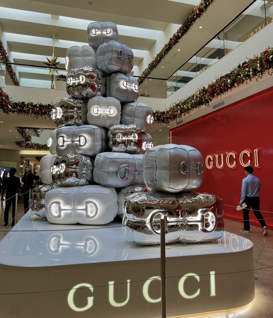 A large, shiny silver and gold sculpture resembling stacked gift bags stands in front of a GUCCI store inside a mall decorated with festive garlands and ornaments. Shoppers walk nearby under bright lighting.