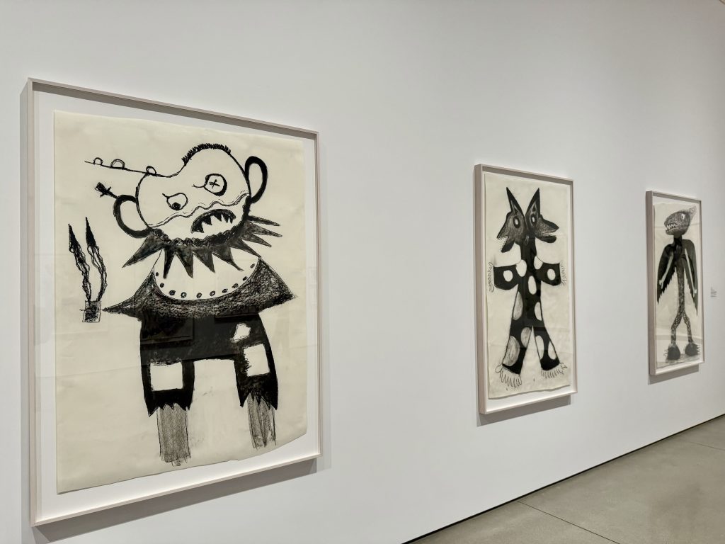 Three large abstract black-and-white drawings in white frames hang on a gallery wall, each depicting imaginative, monstrous figures with bold lines and exaggerated features. The floor and wall are plain and minimalist.