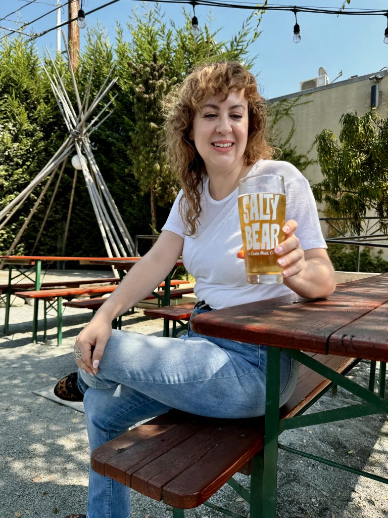 A woman with curly hair and a white t-shirt sits at an outdoor picnic table, smiling and holding a glass of beer labeled SALTY BEAR. There are trees and a teepee structure in the background.