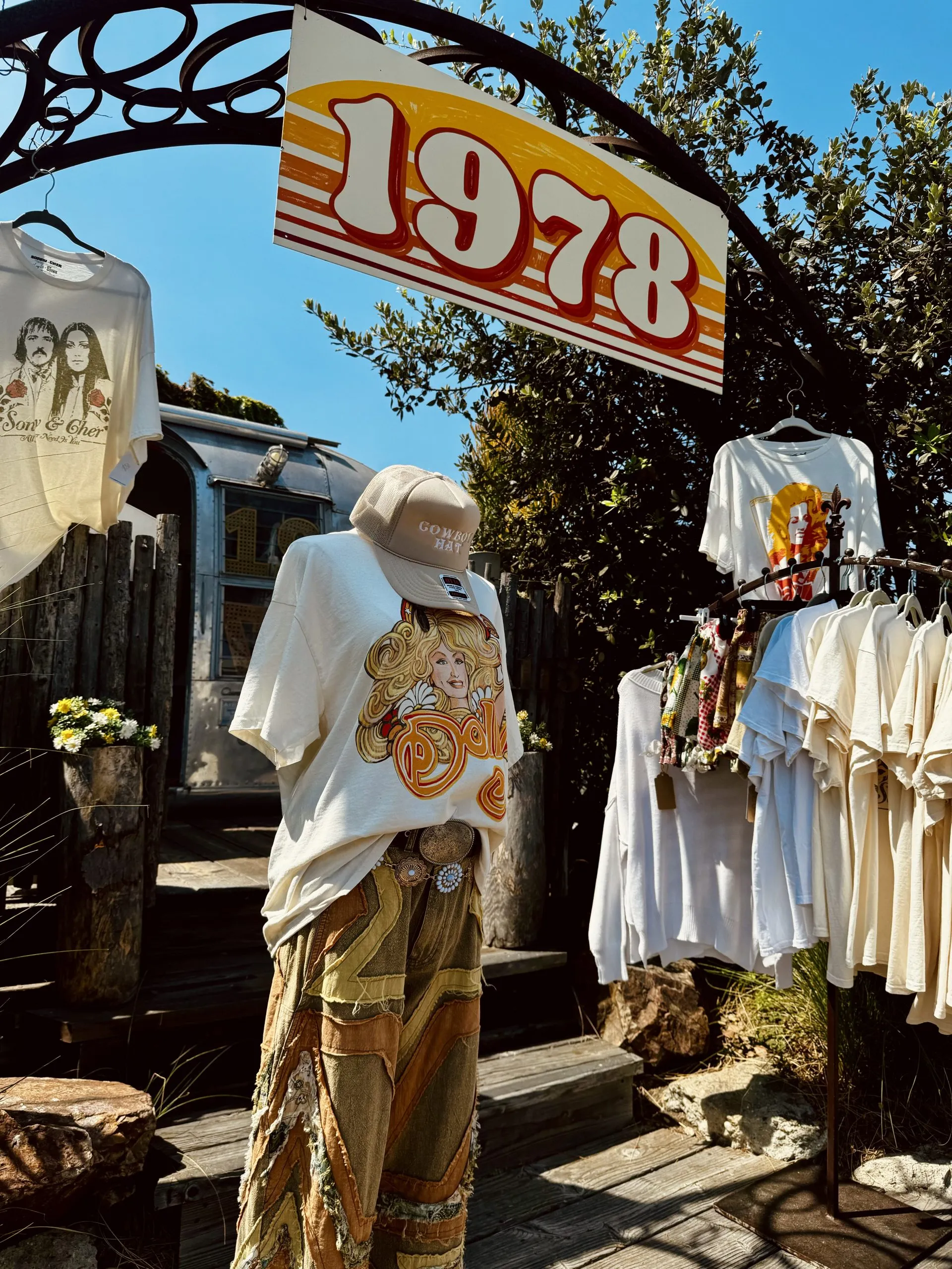 A vintage clothing display outside, featuring a mannequin dressed in retro clothes under a sign reading 1978. T-shirts with 70s-style graphics and other vintage items hang around the setup, with greenery and blue sky in the background.