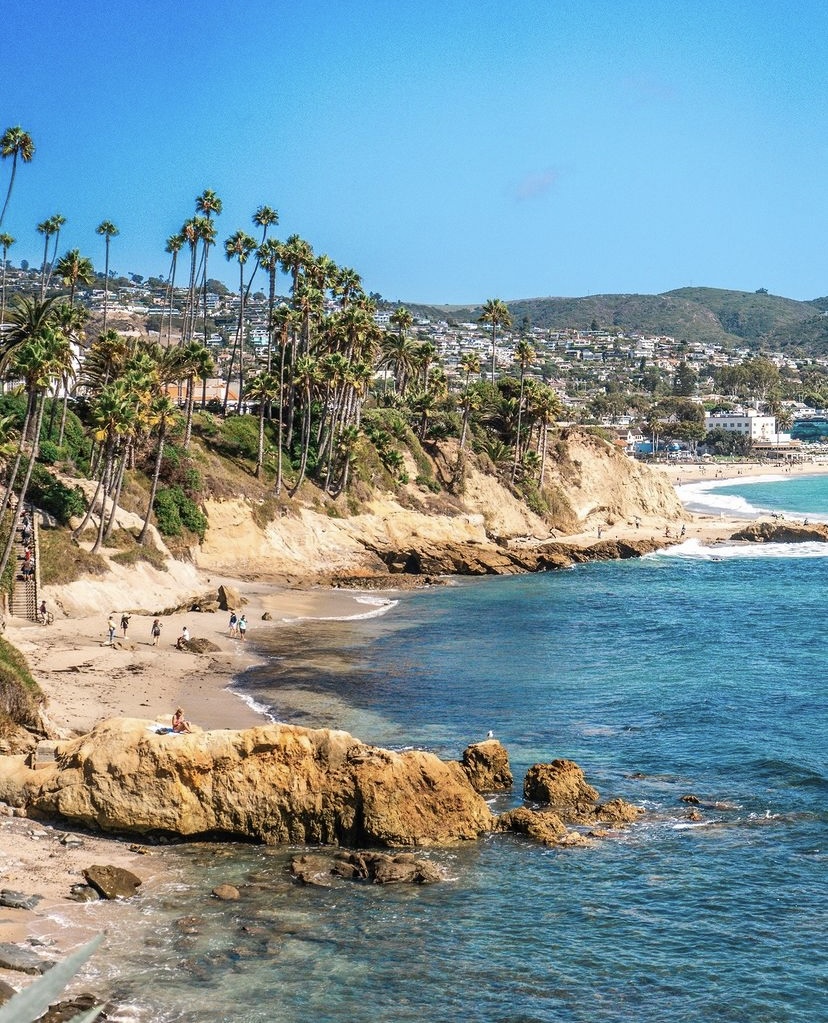 A coastal scene with rocky cliffs, tall palm trees, and sandy beaches bordering clear blue ocean water. People walk along the shore, and houses are visible on the hills in the background under a bright blue sky.