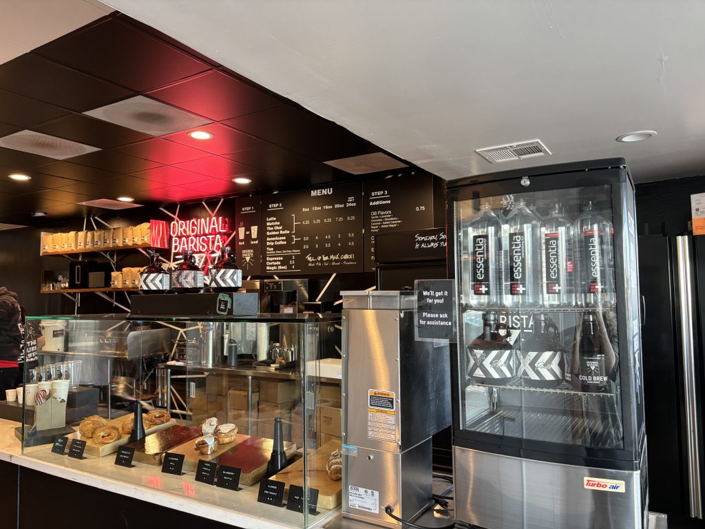 A modern coffee shop counter with pastries on display, a neon sign reading Original Barista, a menu on the wall, coffee machines, and a self-serve water dispenser labeled essentia.